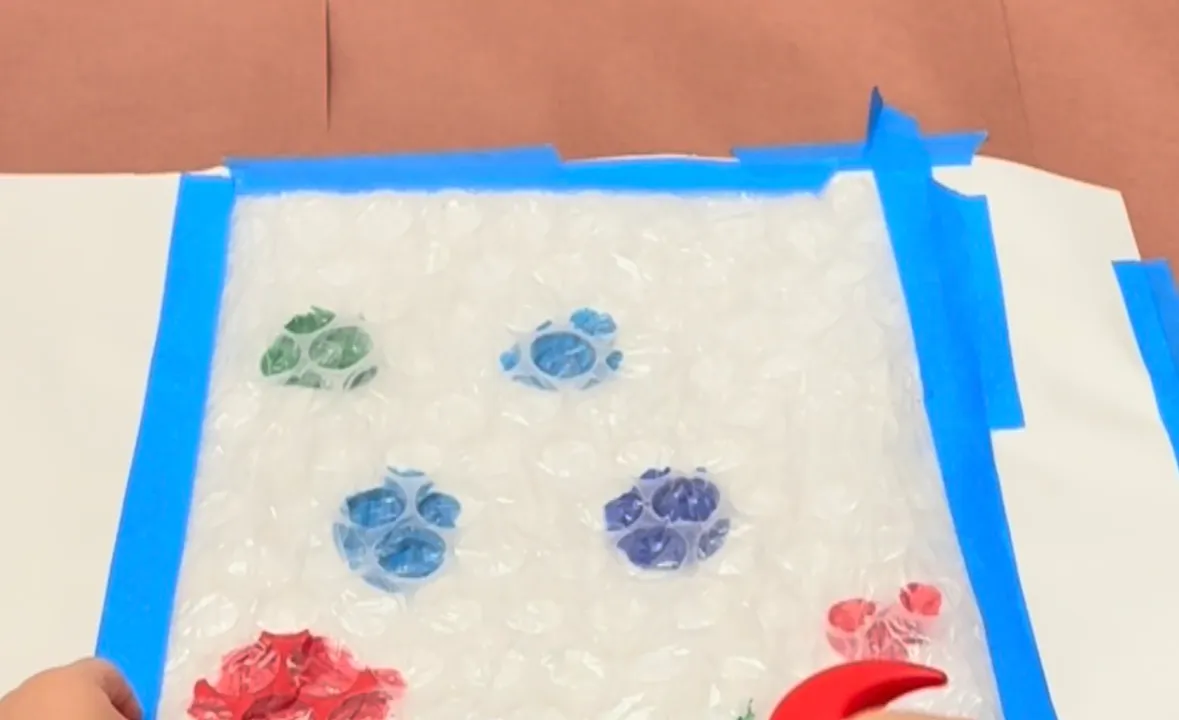 Toddler tapping a toy hammer on bubble wrap taped over blobs of paint on craft paper