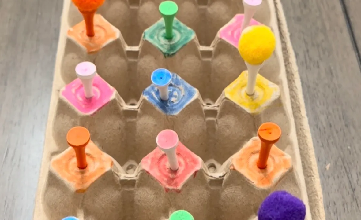 Toddler placing pom poms onto golf tees stuck through the holes of an upside-down egg carton