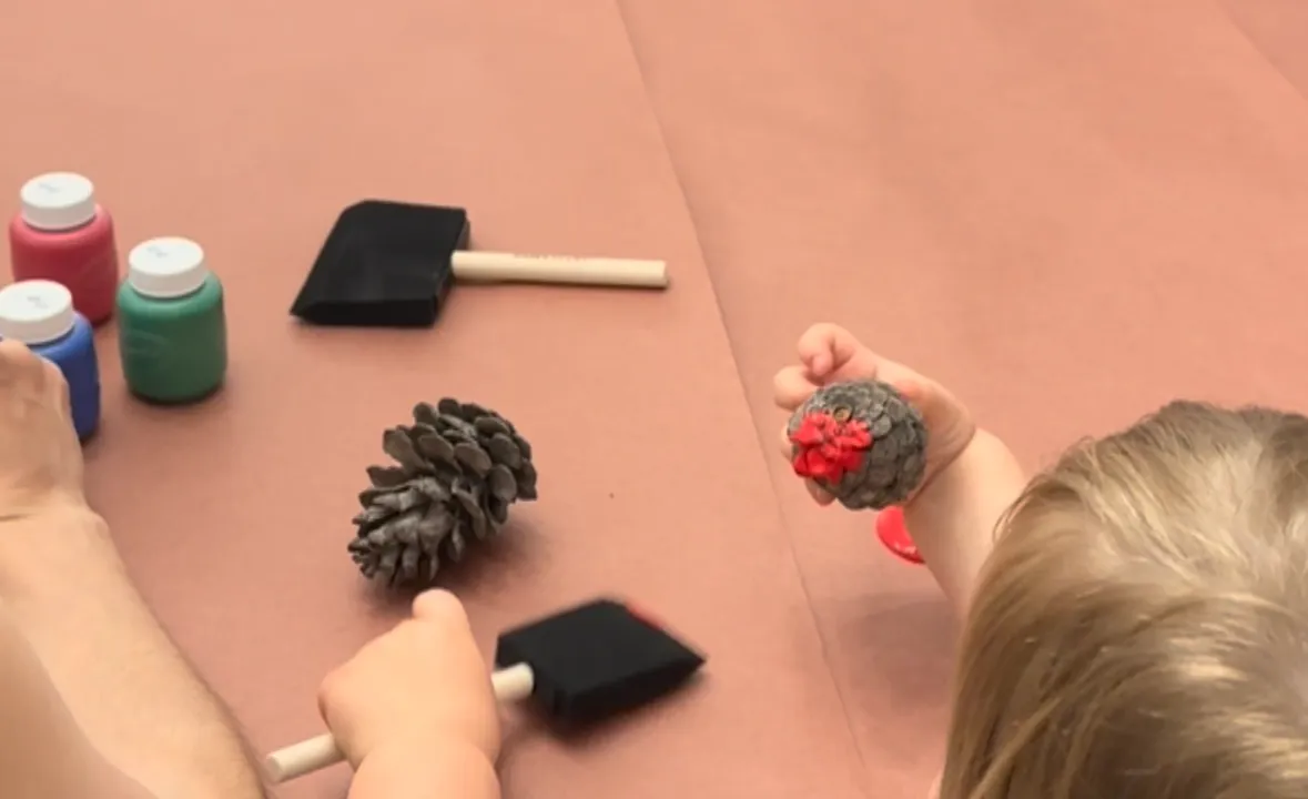Toddler brushing red and yellow paint onto a pine cone on a sheet of paper