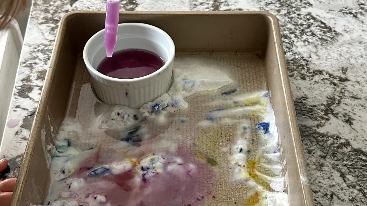 Baking sheet with red, blue, and yellow food coloring covered in white baking soda with a pipette beside it