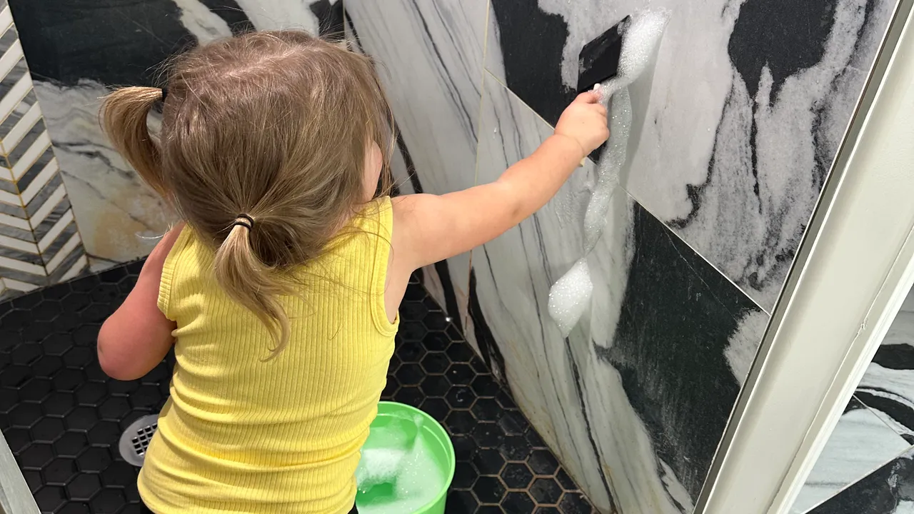 Toddler in a walk-in shower dipping a foam paint brush into a bucket of soapy water and painting bubbles on the tile wall