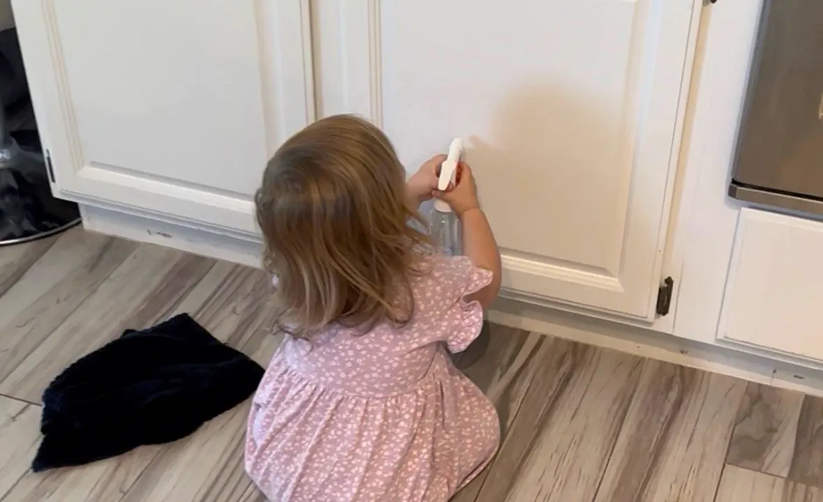 Toddler spraying a kitchen cabinet door with a small spray bottle and wiping it with a cloth