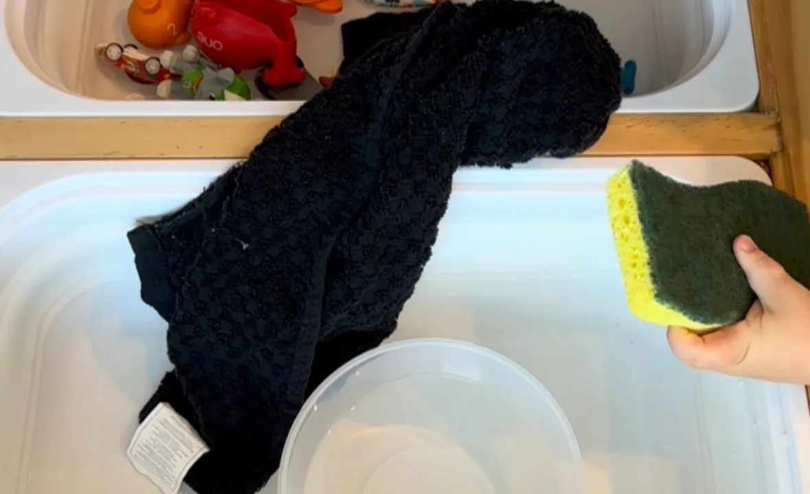 Toddler scrubbing a plastic toy with a sponge over a bowl of soapy water set on a towel