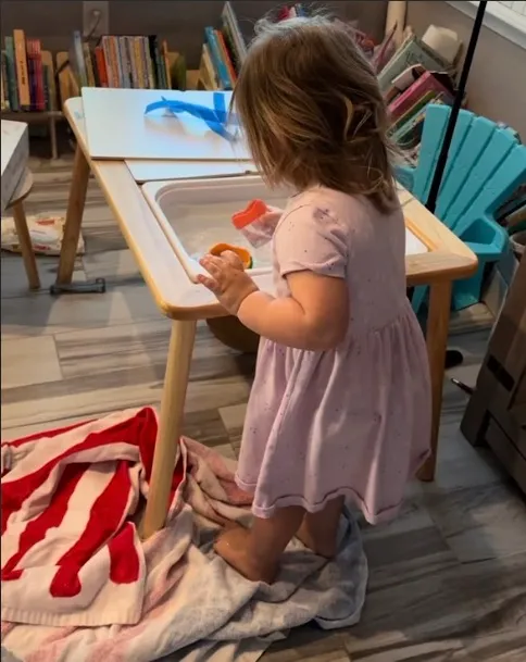 Toddler pouring water from one plastic cup into another over a sensory bin