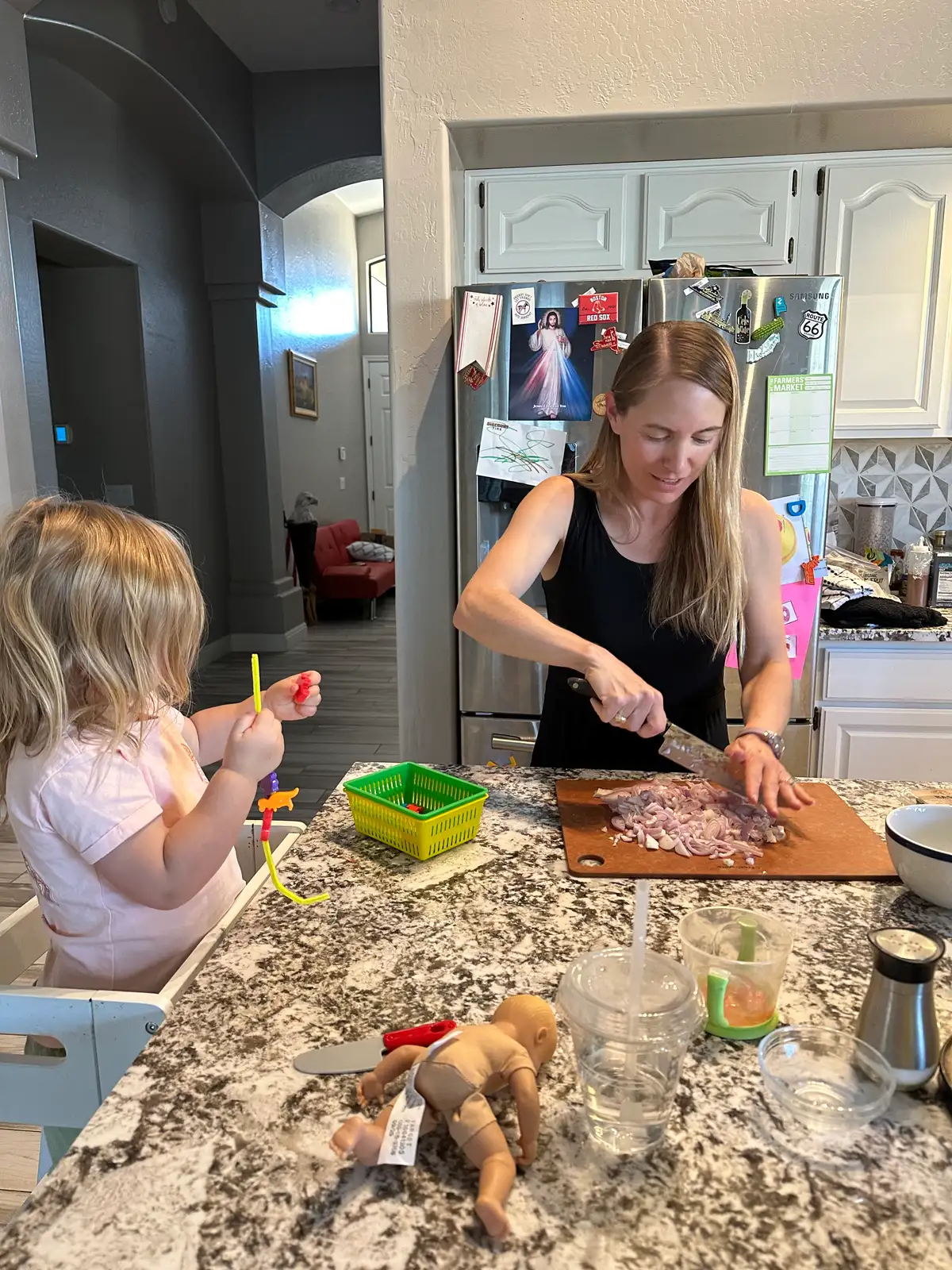 A toddler concentrating on an independent play activity at a low table while a mom cooks in the background.